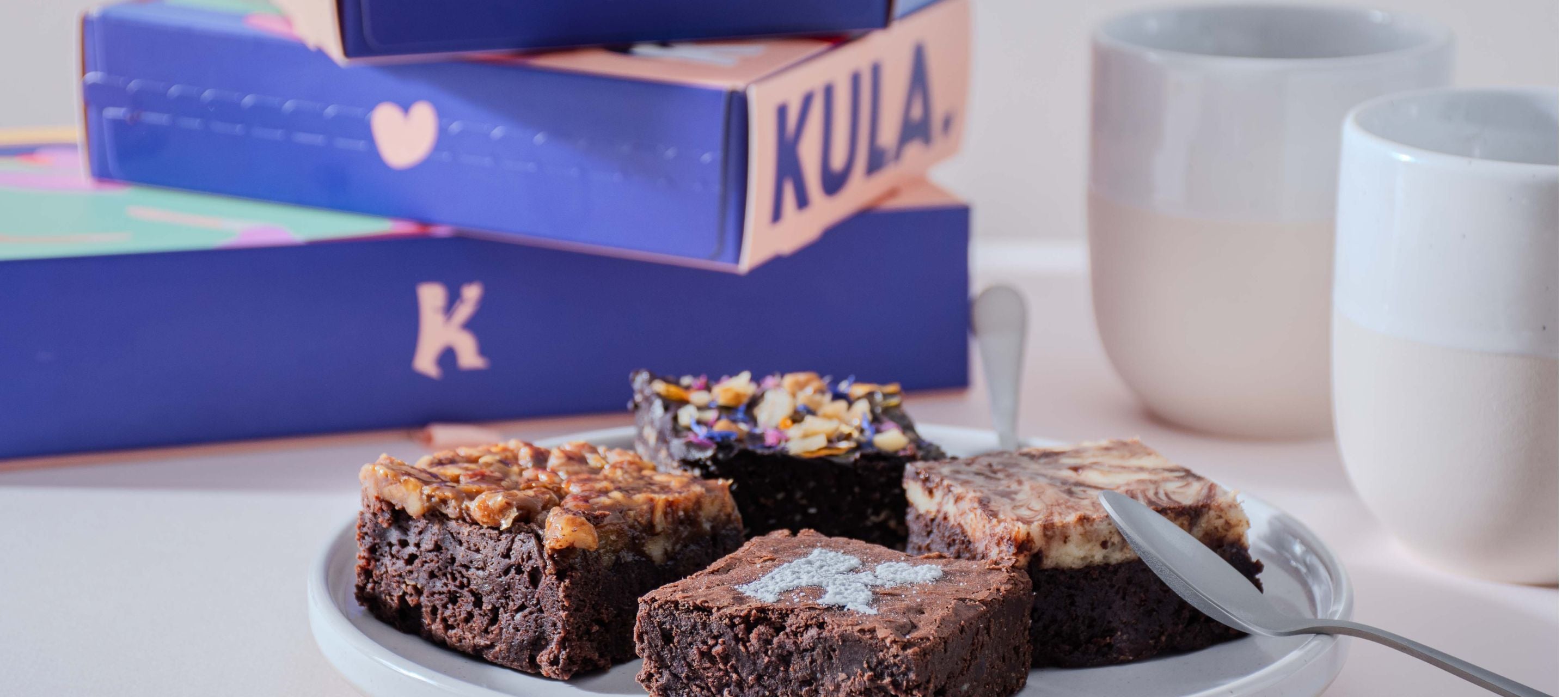 A selection of KULA handcrafted Swiss brownies, including classic, salted caramel, cheesecake, and nutty varieties, elegantly presented on a plate with tea and stacked KULA gift boxes in the background.