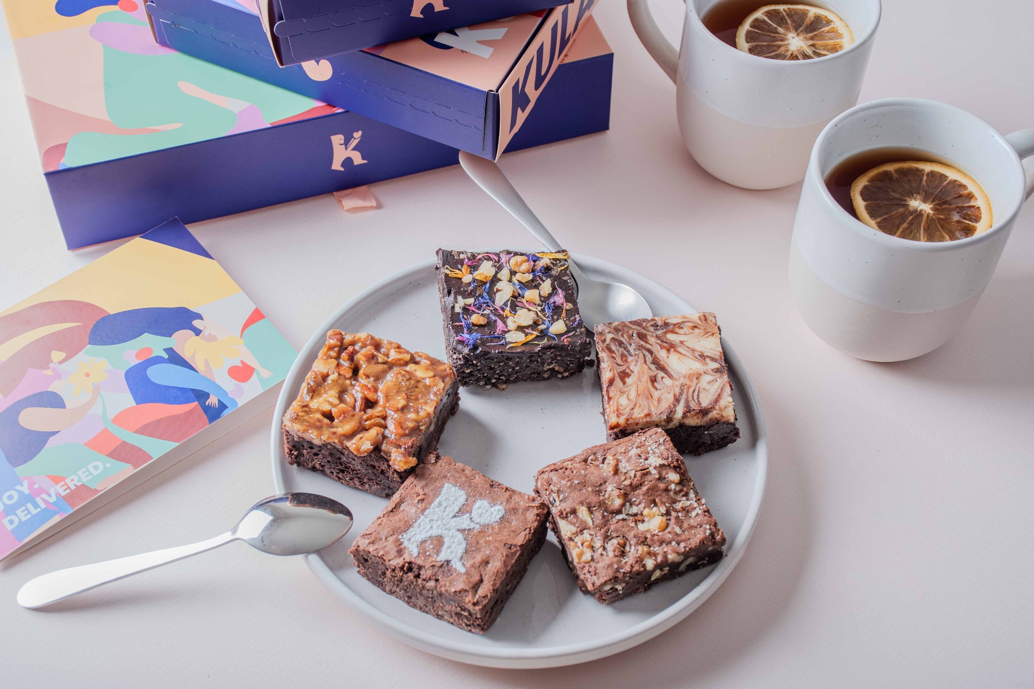 A plate featuring a selection of KULA brownies: Salted Caramel Pecan, Plant-Based Raw Vegan, Cheesecake Swirl, and Classic. The plate is surrounded by KULA gift boxes, colourful cards, and cups of tea showing connection and joy