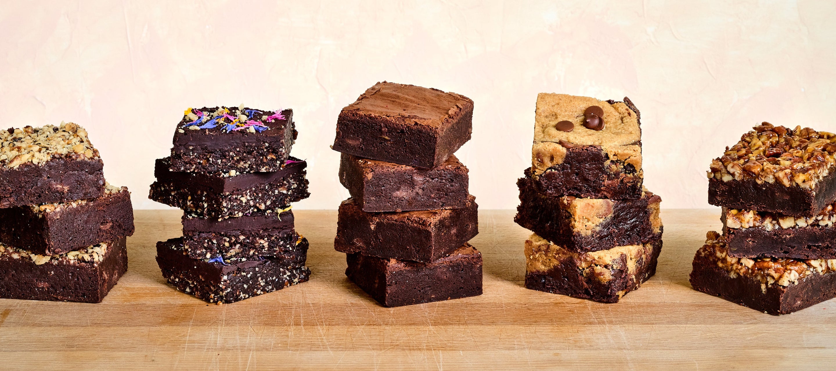 Meet the KULA brownies! An image showing KULA's signature brownies, The Nutty, The Raw, The Classic, The Brookie, The Salted Carmel in stacks, on a wooden board.
