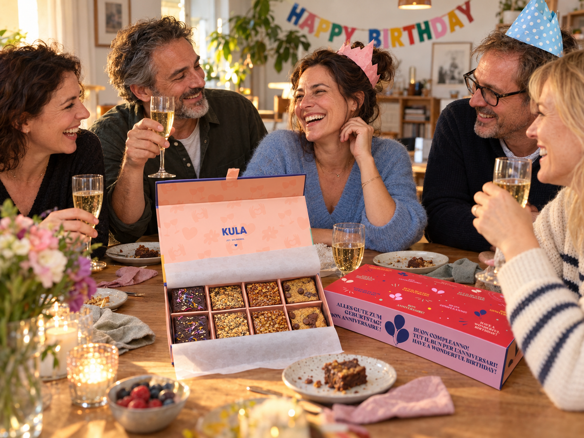Fünf Erwachsene feiern Geburtstag am Tisch mit Sektgläsern, Partyhüten und einer offenen KULA Brownie-Box; die Geburtstagshülle liegt daneben.