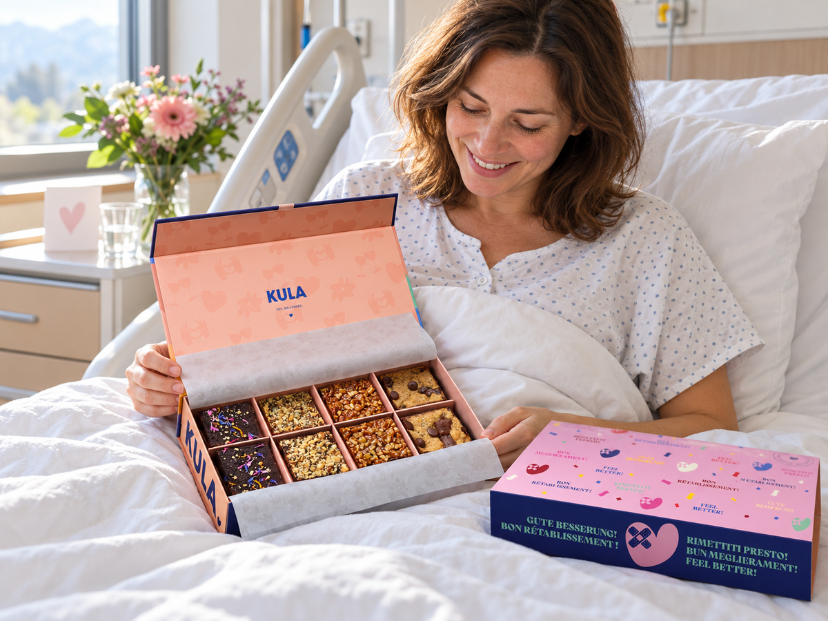 Eine lächelnde Frau im Spitalbett betrachtet eine offene KULA Brownie-Box; daneben liegt die rosa-violette Gute-Besserung-Hülle auf der Bettdecke.