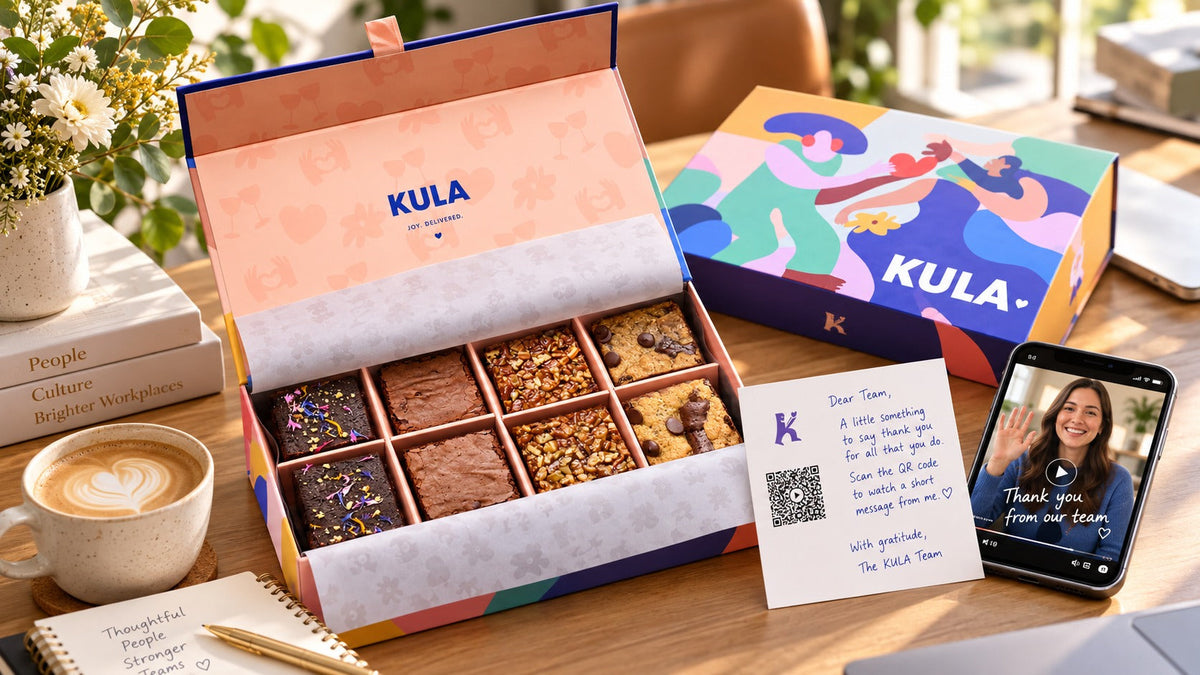 Offene KULA Geschenkbox mit acht Brownies auf einem Bürotisch, dazu eine persönliche Grusskarte mit QR-Code für eine Videobotschaft und ein Smartphone mit Dankesvideo.