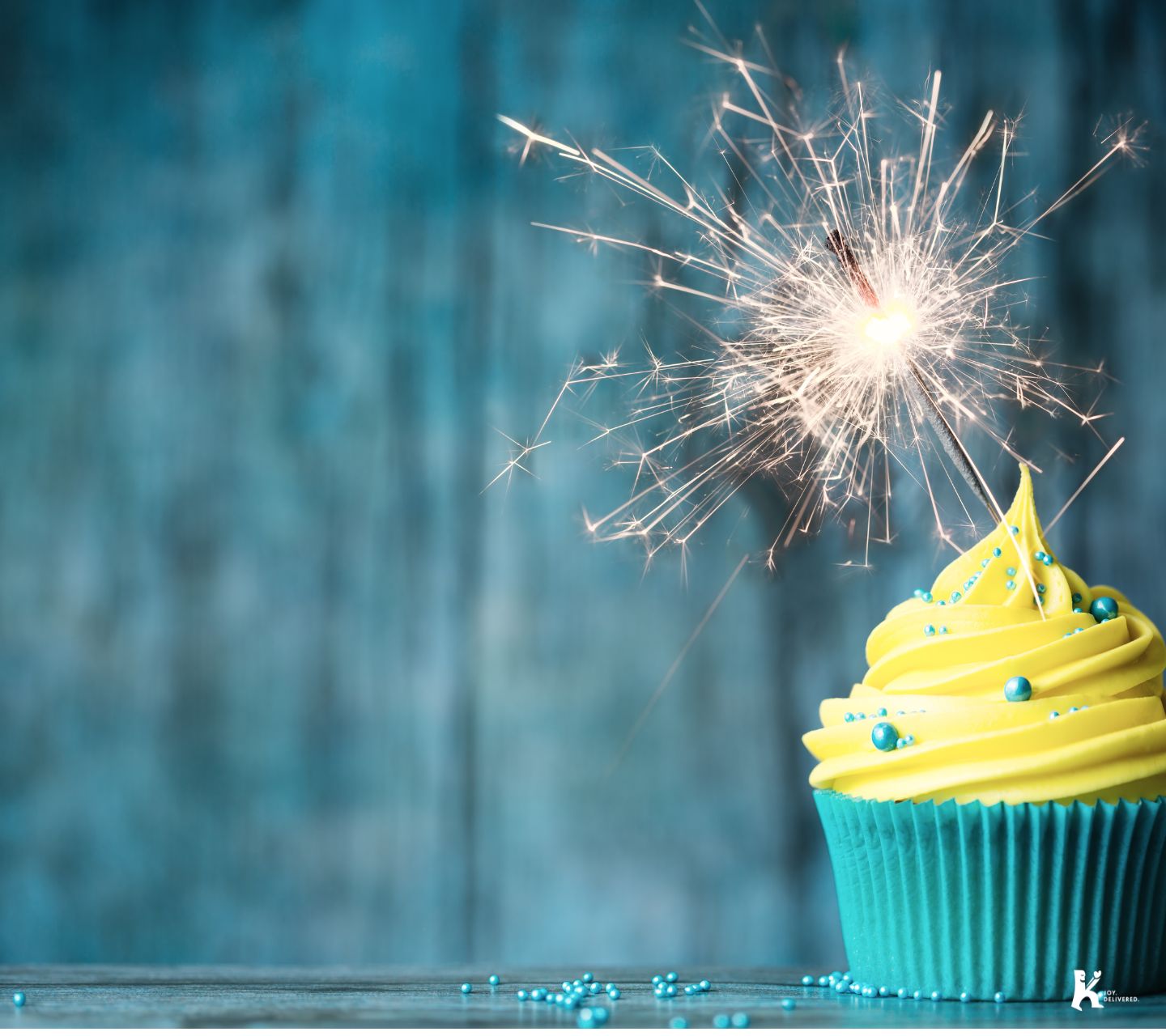 A vibrant cupcake with yellow frosting, blue sprinkles, and a sparkling sparkler on top, symbolizing celebration for the KULA Occasions Collection.