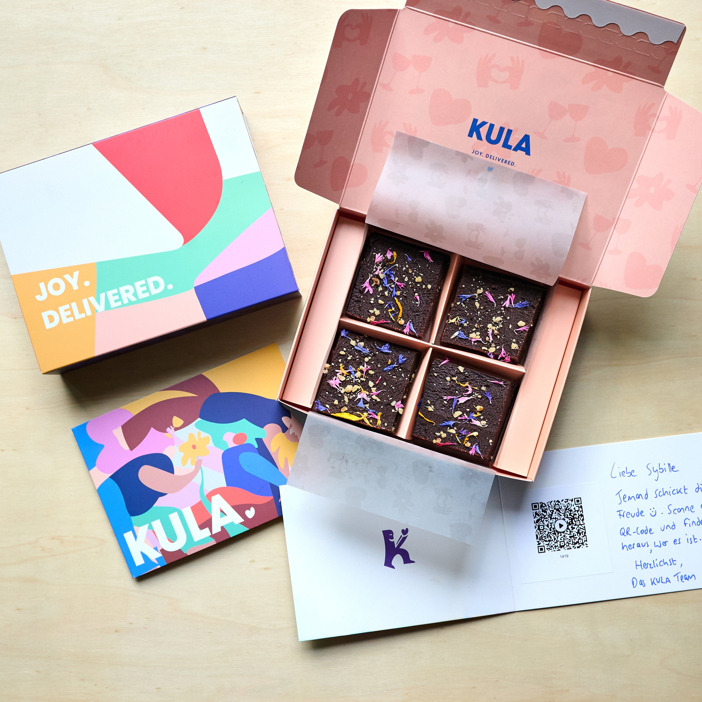 Open KULA gift box with 4 raw vegan brownies topped with colourful edible flowers and nuts. A handwritten card with a QR code for a scannable video message sits beside the box. 