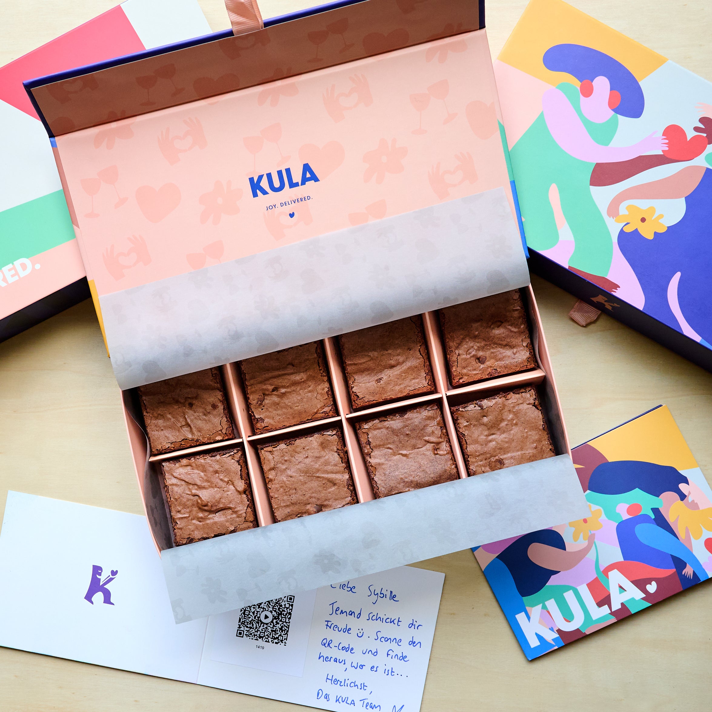 Open KULA 8 box with 8 classic brownies, a handwritten card with scannable personalised video message and a colourful closed KULA box on the side.