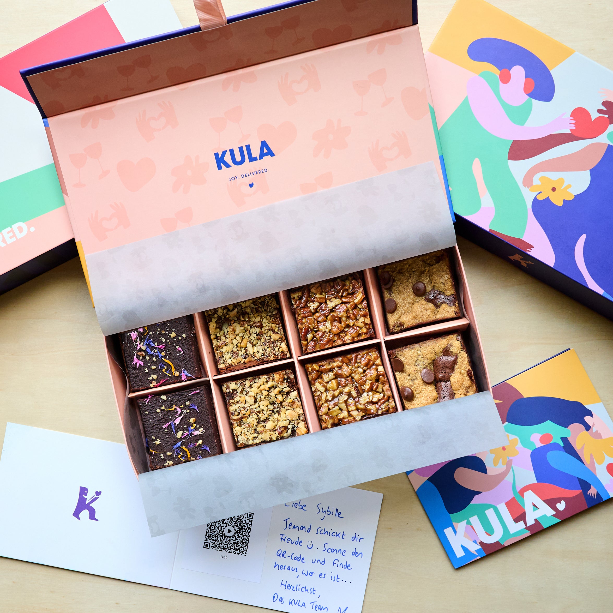 Colourful and vibrant open KULA Gift Box with 8 brownies inside, two Raw, two Nutty, two Salted Caramel and two Brookies. An open handwritten card with scannable personal video message is beside the box. 