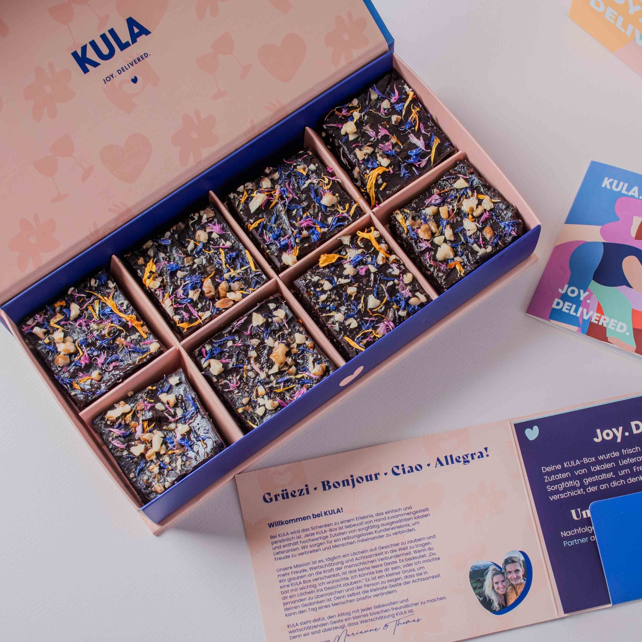 An open KULA gift box featuring eight squares of plant-based raw vegan brownies, beautifully decorated with vibrant edible flowers and nuts. Accompanied by a handwritten card and KULA’s colourful and inviting packaging, perfect for thoughtful gifting and sharing.