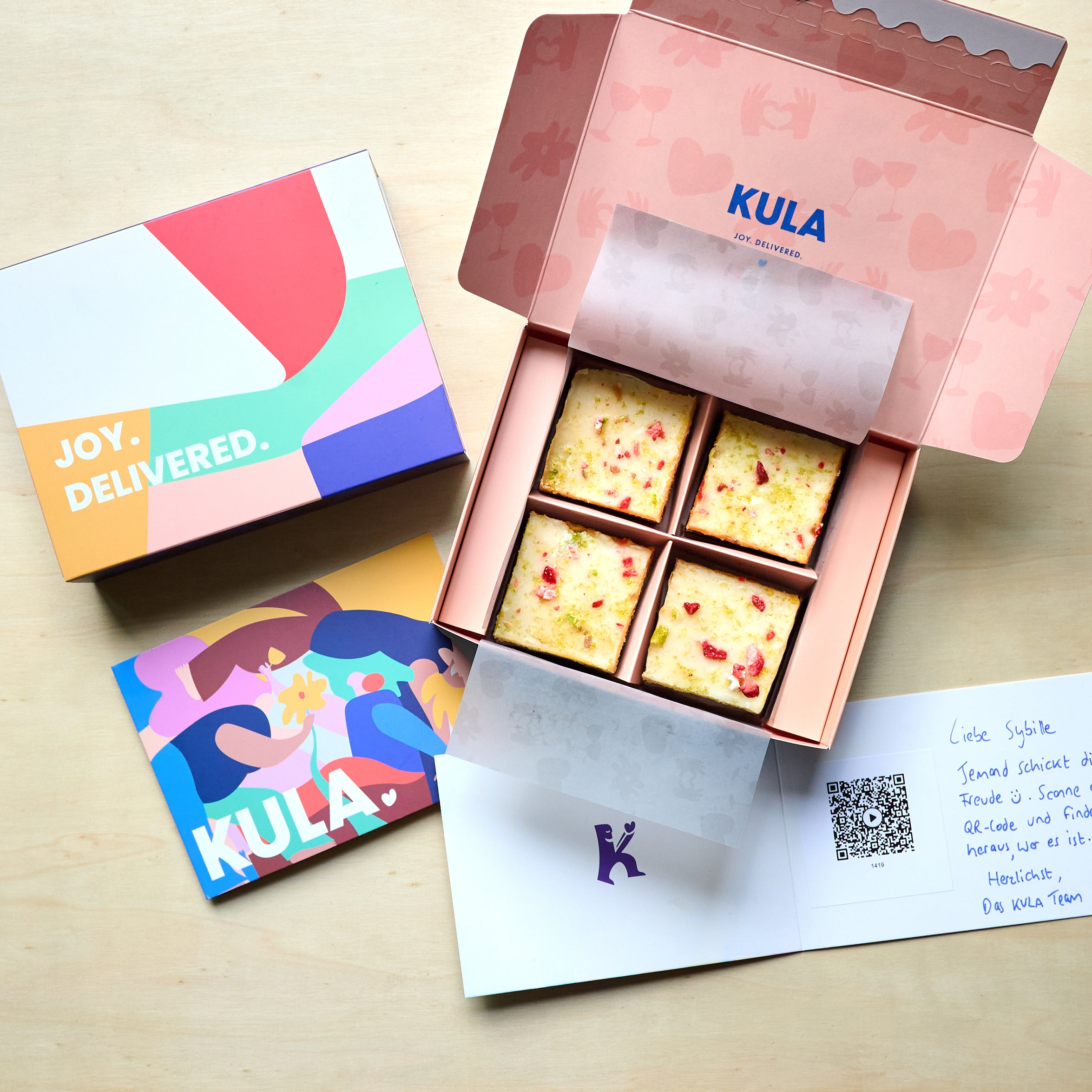 Open KULA gift box with 4 lemon and lime blondies, topped with freeze-dried strawberry pieces and lemon and lime zest. A handwritten card with a scannable QR code for a personal video message sits beside the box. 