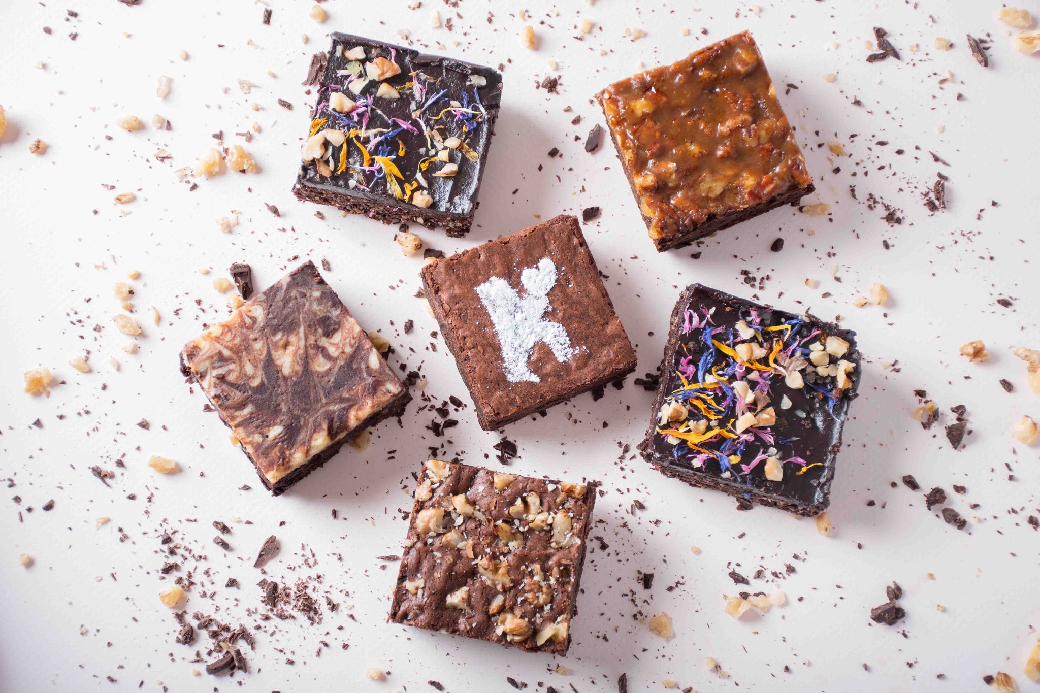 An artistic flat-lay of six KULA brownies featuring distinct flavours: Plant-Based Raw Vegan, Salted Caramel Pecan, Cheesecake Swirl, Nutty Walnut, Classic with powdered sugar 'K' design, and more. Scattered chocolate shavings and nuts add a creative touch