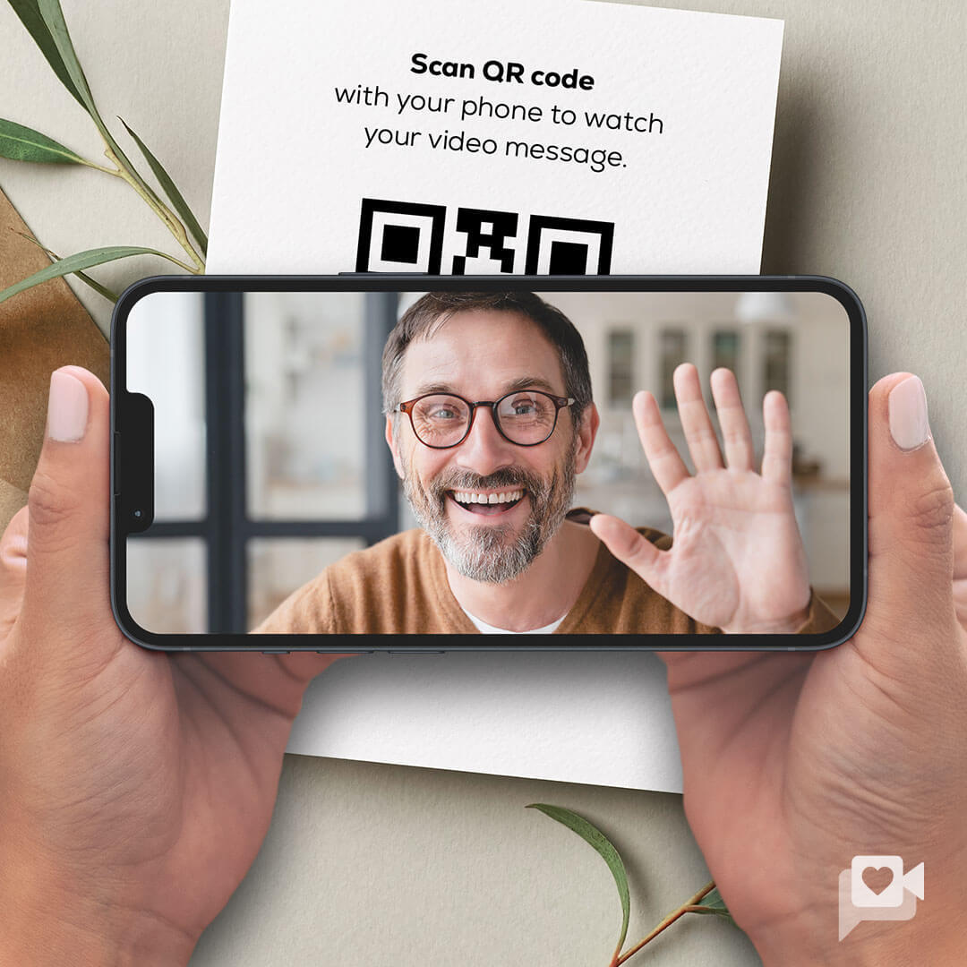 A person holding a phone displaying a smiling man waving, with a QR code in the background. Scan the code to watch a heartfelt KULA personal video message included with your brownie gift.