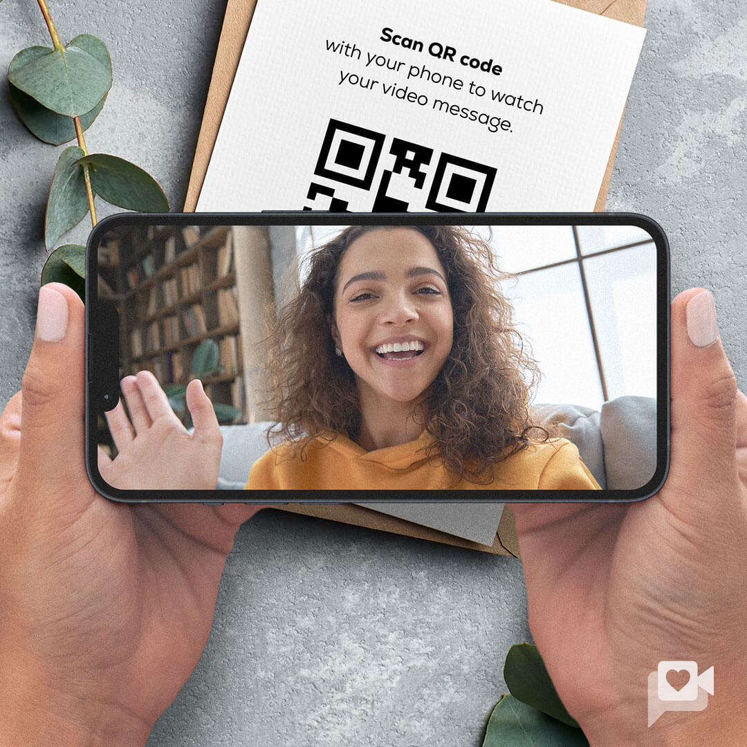 A person holding a phone displaying a smiling woman waving, with a QR code in the background. Scan the code to watch a personalized KULA video message included with your brownie gift.