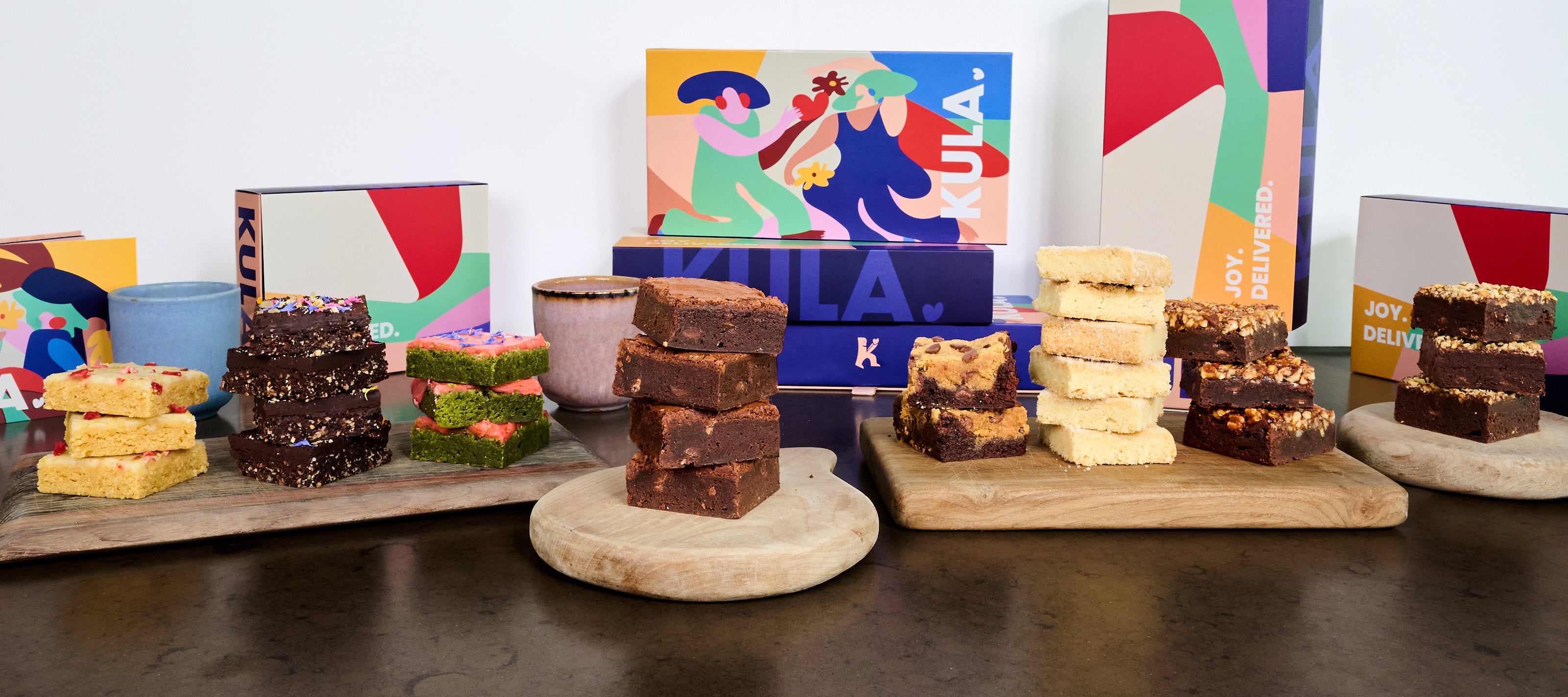 Wide image showing all KULA brownie flavours in stacks in front of colourful KULA boxes.